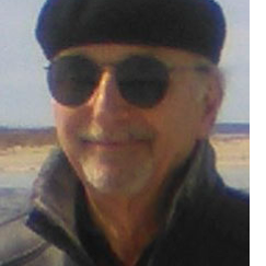 Headshot of a man wearing a Basque-style beret, round sunglasses and winter coat standing on a beach somewhere on the north shore of Long Island.